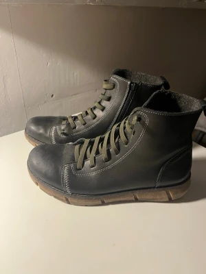 Svarta kängor med dragkedja och snörning - Svarta kängor i skinn med gröna snören, grov sula i beige och vit kontrastsöm. Insidan är fodrad med grått tyg för extra värme. Dragkedja på sidan gör dem enkla att ta på och av. Perfekta för dig som vill ha en stilren och robust look.