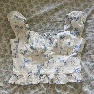 Vit blommig korsettopp från Hollister - Supersöt vit korsettopp från Hollister med ljusblått blommönster, volangkant nertill och spetsdetaljer. Toppen har korta puffärmar, snörning framtill och croppad passform. Perfekt till sommaren och ger en romantisk vibe till din outfit. 🩵🤍 använd ett fåtal gånger. Köptes för 419kr på Zalando och säljer då den inte kommer till användning