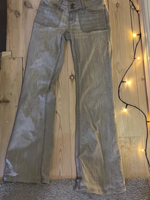 Gråa bootcut jeans med låg midja  - Säljer ett par ljusgrå jeans med bootcut passform och stora fickor både fram och bak. Passar någon som är runt 160 cm  Jeansen har dubbla knappar i midjan och coola ficklock bak med knappdetaljer. Perfekta för dig som gillar en avslappnad men ändå snygg stil. 