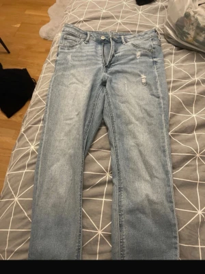 Ljusblå jeans med slitna detaljer - Säljer ett par ljusblå jeans med raka ben och slitna detaljer vid fickor och benslut. Jeansen har klassisk femficksdesign och råa, fransiga kanter längst ner. 