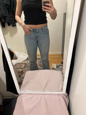 Ljusblå bootcut jeans med slitningar - Säljer ett par ljusblå jeans med bootcut passform och slitna detaljer vid knäna. Jeansen har klassisk femficksdesign, låg midja och är tillverkade i mjukt denimtyg. Perfekta för en avslappnad och trendig look.