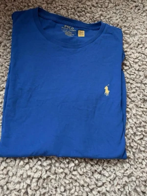 Blå t-shirt från Polo Ralph Lauren - Snygg blå t-shirt från Polo Ralph Lauren med klassisk gul broderad logga på bröstet. Enkel och stilren design med rund halsringning och korta ärmar. Tillverkad i mjuk bomull som känns skön mot huden.