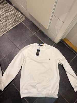 Vit sweatshirt från Polo Ralph Lauren - Säljer en clean vit sweatshirt från Polo Ralph Lauren med klassisk blå logga på bröstet. Tröjan har rund halsringning, ribbade muddar och är i mjuk bomull. Perfekt basic som funkar till allt och ger en fräsch look. Har aldrig använts!