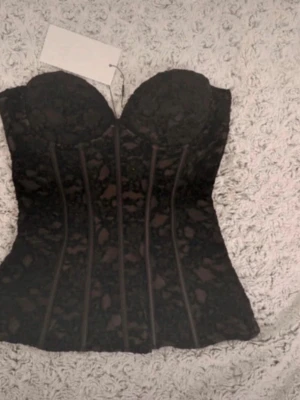Svart spetskorsett med struktur - Säljer en svart korsett med vacker spets och markerade sömmar framtill som ger en snygg siluett. Toppen har bygelkupor och är axelbandslös, perfekt för att styla till olika looks. Materialet känns lyxigt och detaljerna ger en edgy vibe.