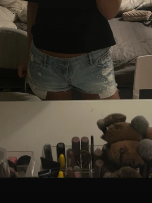 Ljusblå jeansshorts Hollister W28 - Säljer ett par ljusblå slitna jeansshorts från Hollister med råa kanter, slitningar och coola nitar och pärldetaljer framtill. Klassisk femficksmodell med låg midja och snygga broderade bakfickor. Perfekta till sommaren!