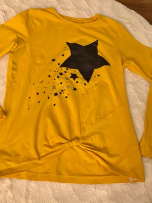 Gul långärmad topp med stjärntryck - Cool gul långärmad topp från Name It i ekologisk bomull. Framsidan har ett stort svart stjärntryck med mindre stjärnor i svart och silver, samt en snygg knutdetalj nedtill. Perfekt för dig som gillar färg och vill sticka ut lite extra.