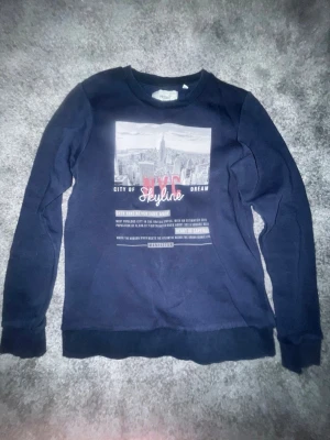 Mörkblå sweatshirt från Produkt - Mörkblå sweatshirt från Produkt med tryck av New Yorks skyline och texten 'NYC Skyline' på bröstet. Tröjan har rund halsringning och långa ärmar. Perfekt för dig som gillar streetstyle och vill ha en bekväm och snygg tröja.
