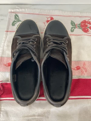 Bruna lanvins uk5 - Snygga sneakers i mörkbrun mocka med svart, glansig tå och vita sulor. Skorna har snörning med metallöljetter och en klassisk låg profil. Insidan är fodrad i svart skinn för extra komfort. Perfekta för dig som gillar stilrena detaljer. Uk5 vilket är str 39 men sitter mer som 40!