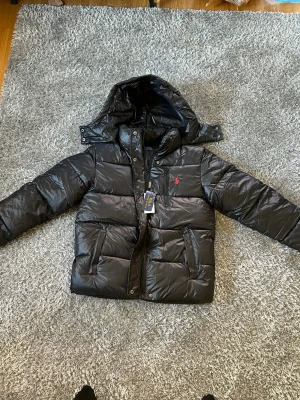 Svart pufferjacka från Polo Ralph Lauren - Säljer en svart pufferjacka från Polo Ralph Lauren med huva och dragkedja. Jackan har en röd broderad logga på bröstet och stora fickor framtill. Materialet är glansigt och känns riktigt modernt. Perfekt för kalla dagar när du vill hålla stilen.