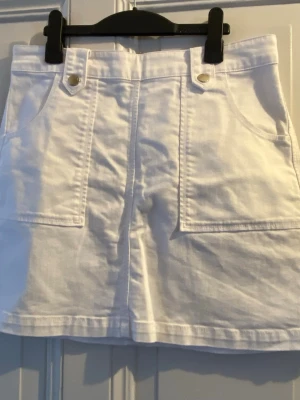 Vit kjol från Bondelid - Säljer en vit kortkjol från Bondelid. Kjolen har stora fickor fram, dekorativa sömmar och en dragkedja bak. Snygga silverfärgade knappar framtill ger en clean och fräsch look. Perfekt till sommaren och enkel att matcha. Köpt på MQ.