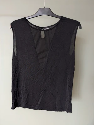 Svart mesh ärmlös topp KappAhl - Säljer en svart ärmlös topp från KappAhl med meshdetaljer framtill och baktill. Toppen har en djup v-ringning i mesh och en liten droppformad öppning vid halsen. Materialet är tunt och lätt, perfekt för att styla lager på lager.
