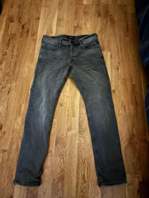 Grå jeans från Jack & Jones, 29/30 - Säljer ett par grå jeans från Jack & Jones i storlek 29/30. Jeansen har klassisk femficksdesign, raka ben och diskret tvättad look. Tillverkade i mjukt bomullsmaterial med en snygg, lite sliten finish. Perfekta till sneakers och hoodie.