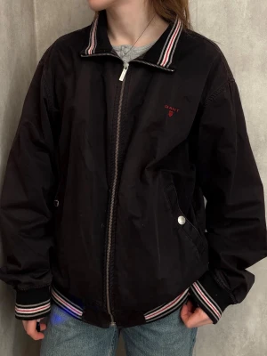 GANT - jacka  - Säljer en brun jacka från GANT med röd logo på bröstet. Randiga detaljer vid kragen, ärmarna och jackkanten. Har själv använt den som en oversized vårjacka. Diskret och stilren ;)