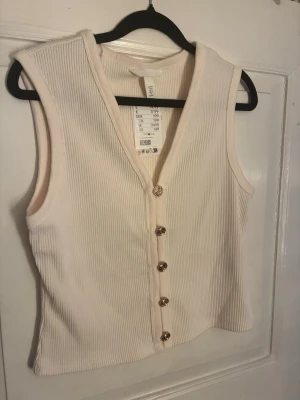 Ribbad beige väst med guldfärgade knappar - Säljer en ribbad beige väst från H&M i storlek M. Västen har v-ringning och fem dekorativa guldfärgade knappar framtill. Materialet är mjukt och stretchigt, perfekt för att styla lager på lager. Enkel och stilren design som ger en fräsch look. Helt ny med prislapp kvar.