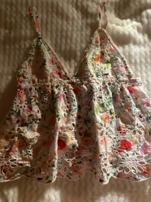 Blommig broderad linnetopp Gina Tricot - Superfin blommig linnetopp från Gina Tricot i storlek 40. Toppen har tunna axelband, v-ringning och är broderad med hålmönster och färgglada blommor i rosa, rött, grönt och orange. Perfekt för varma dagar och ger en romantisk vibe.