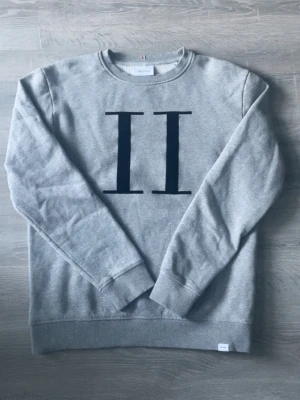 Grå sweatshirt Les Deux  - Säljer en stilren grå sweatshirt från Les Deux i storlek M. Tröjan har ett stort svart II-tryck på bröstet, rund halsringning och ribbade muddar. Tillverkad i mjuk bomullsblandning som är perfekt för både höst och vår.