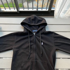 Svart hoodie från Polo Ralph Lauren - Svart hoodie från Polo Ralph Lauren med dragkedja och huva. Klassisk design med broderad logga på bröstet och känguruficka framtill. Tillverkad i mjuk bomull, perfekt för chill dagar eller när du vill ha en clean streetstyle. Storlek M men passar S.