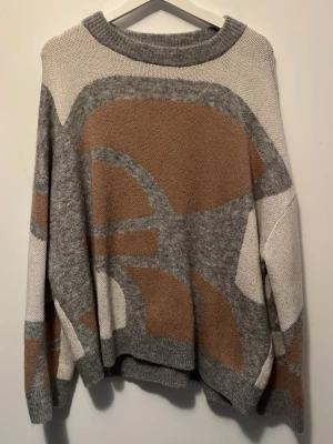 Beige och grå stickad tröja med mönster - Säljer en oversized stickad tröja i beige, grått och brunt med abstrakt mönster. Tröjan har rund halsringning och långa ärmar. Perfekt för dig som gillar en chill och unik stil.