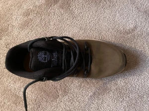 Timberland boots i olivgrönt skinn - Säljer ett par snygga Timberland boots i olivgrönt/brunt skinn med svarta meshdetaljer och svart snörning. Skorna har en robust vit och svart sula med grovt mönster för extra grepp. Perfekta för dig som vill ha varma men snygga skor