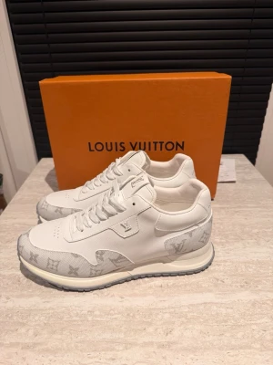Vita Louis Vuitton sneakers med monogram - Louis Vuitton sneakers i vitt skinn och textil med ikoniskt monogrammönster i grått på tå och häl. Skorna har rund tå, snörning och en rejäl grå sula med LV-logga undertill. Perfekta för dig som vill ha en clean och lyxig look.