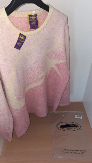 Ljusrosa stickad Corteiz tröja  - Ljusrosa stickad tröja från Corteiz med ett stort stjärnmotiv i vitt framtill. Tröjan har rund halsringning, ribbade muddar och lång ärm. Perfekt för dig som vill ha en mysig och trendig look till hösten eller vintern.