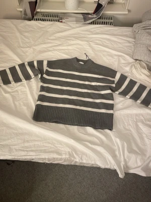 Gråvit randig stickad tröja H&M XS - Mysig stickad tröja från H&M i grått och vitt med breda ränder. Tröjan har rund hals, ribbade muddar och lång ärm. Perfekt till jeans för en chill look. Passar dig som gillar en normal passform och vill ha något enkelt men ändå snyggt.