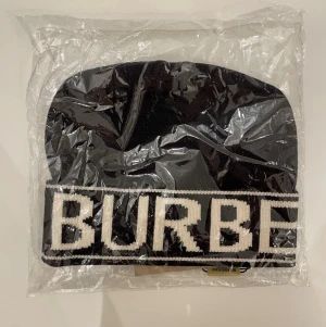 Svart Burberry mössa med logga - Säljer en svart mössa från Burberry med stor vit logga runt om. Mössan är ribbstickad och har en bred kant med tydlig text. Perfekt accessoar för dig som vill ha en stilren och lyxig look.