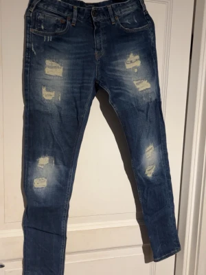 Blå slitna jeans från Scotch & Soda - Säljer ett par blå jeans från Scotch & Soda med snygga slitningar och distressed detaljer framtill. Jeansen har klassisk femficksdesign, normal passform och är tillverkade i jeansmaterial. Hör av er vid funderingar✌️