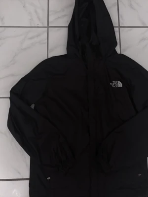 Svart vindjacka från The North Face - Säljer en svart vindjacka från The North Face med huva och diskret logga på bröstet. Jackan är tillverkad i ett lätt och tåligt syntetmaterial som är både vattentätt och andas. Perfekt för utomhusaktiviteter och har en clean, sportig look. Den är i storlek S men funkar även som XS. 
