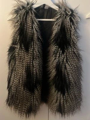 Pälsväst - Säljer en fluffig och randig pälsväst från H&M Divided i storlek 38. Västen har svart och grå ton med coolt mönster och är tillverkad i syntetpäls. Perfekt att slänga över en hoodie eller tröja för extra edge. Fodrad insida för skön känsla.