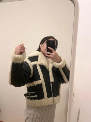 Svart teddyjacka med fuskpälsdetaljer - Säljer en svart teddyjacka från Gina Tricot med krämvit fuskpäls längs krage, ärmslut och kanter. Jackan har en cool, boxig passform, dragkedja framtill och bältesdetaljer i sidorna. Perfekt för dig som vill sticka ut i vinter med en trendig look.Knappt använd,som ny! 