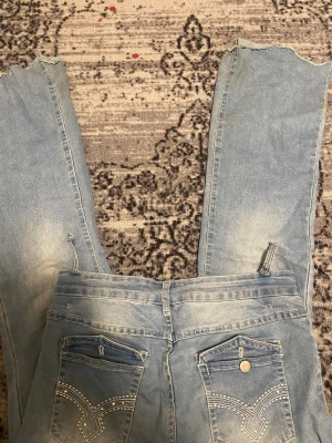 Ljusblå bootcut jeans med råa kanter - Säljer ett par ljusblå jeans med bootcut-modell och råa, slitna kanter nertill. Jeansen har klassiska bakfickor med dekorativa sömmar och knappdetalj. Perfekt för dig som gillar en avslappnad och trendig look.