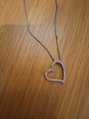 Vintage Halsband med hjärthänge i silverfärg - Säljer ett smalt halsband i silverfärg med ett hjärthänge dekorerat med små glittrande stenar. Kedjan är tunn och smidig, och hjärtat har en öppen design som ger ett stilrent och sött intryck. Perfekt accessoar för att lyfta vilken outfit som helst.
