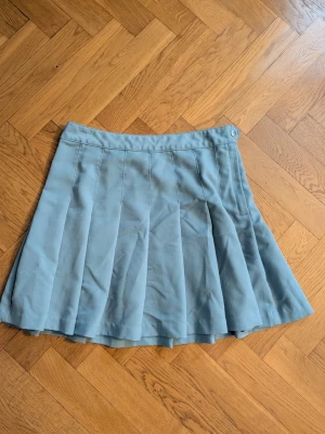 Ljusblå plisserad kortkjol - Säljer en ljusblå plisserad kortkjol med snygga veck och en diskret dragkedja i sidan. Kjolen har en knappdetalj i midjan och är gjord i ett mjukt bomullsmaterial som känns bekvämt. Perfekt för dig som gillar stilrena och enkla plagg.