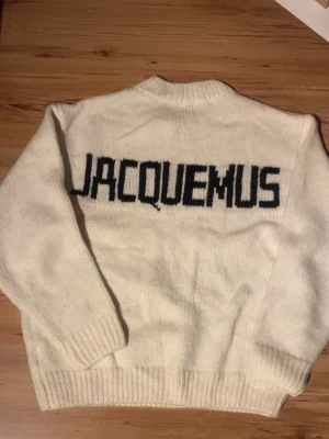 Vit stickad tröja från Jacquemus - Säljer en vit stickad tröja från Jacquemus med svart logotyp på ryggen. Tröjan har rund hals, ribbade muddar och nederkant samt långa ärmar. Perfekt för dig som gillar stilrena och trendiga plagg med tydlig branding.