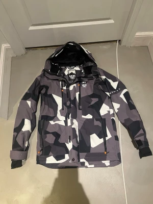 Camouflage skidjacka från Montec - Säljer en snygg skidjacka från Montec med grå, svart och vit camouflage-mönster. Jackan har huva, dragkedja framtill, flera fickor och en ficka i armen för liftkort. 