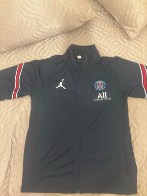 PSG x Jordan träningsjacka marinblå - Marinblå träningsjacka från PSG x Jordan med dragkedja, vita och röda ränder på ärmarna samt PSG-logga och Jumpman-logga på bröstet. Jackan har hög krage och fickor framtill. Perfekt för dig som gillar sportig stil.