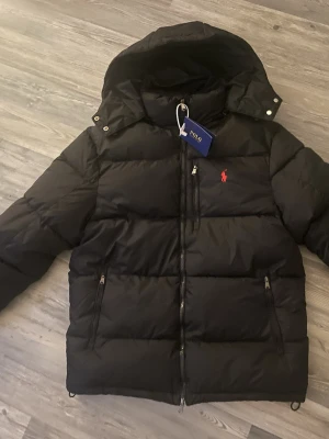 Svart pufferjacka från Polo Ralph Lauren - Säljer en svart pufferjacka från Polo Ralph Lauren med huva och dragkedja. Jackan har det klassiska röda Polo-logot broderat på bröstet och flera praktiska fickor med dragkedja. Perfekt för kalla dagar och riktigt snygg till streetwear-stilen.