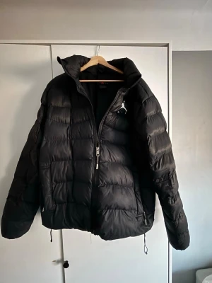 Jordan svart pufferjacka loose fit - Säljer en svart pufferjacka från Jordan i loose fit. Jackan har hög krage, dragkedja framtill och två sidofickor med dragkedja. Klassisk Jordan-logga på bröstet och justerbar nederkant med snörning. Perfekt för kalla dagar och riktigt snygg streetstil.