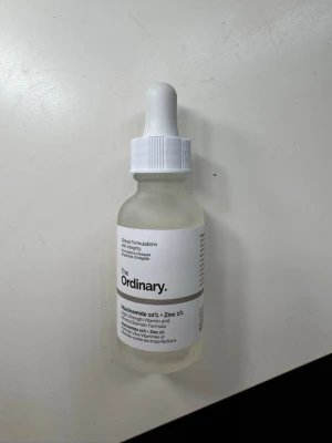 The Ordinary Niacinamide 10% + Zinc 1% - Transparent ansiktsserum i glasflaska med vit pipett från The Ordinary. Innehåller niacinamid 10% och zink 1% för att balansera huden och minska orenheter. Enkel, stilren design med vit etikett och svart text.