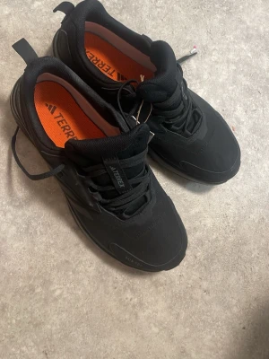 Svarta Adidas Terrex sneakers - Säljer ett par svarta Adidas Terrex sneakers med orange innersula och robust design. Skorna har snörning, rund tå och slitstark ovandel i syntetmaterial. Perfekta för utomhusaktiviteter och äventyr. Märket syns tydligt på plösen och innersulan. Helt ny