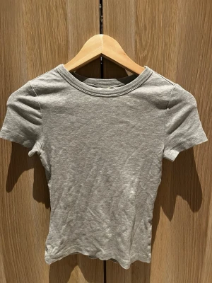 Grå ribbad t-shirt från H&M XS - En enkel och stilren grå t-shirt från H&M i storlek XS. T-shirten är ribbad, har rund halsringning och korta ärmar. Tillverkad i mjuk bomull som känns skön mot huden. Perfekt basplagg till alla outfits.