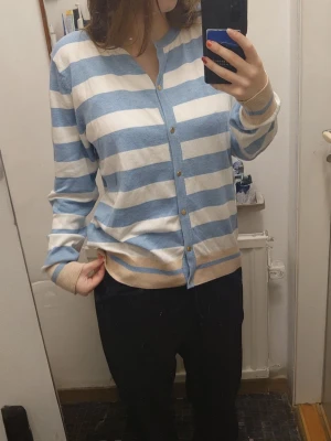 Randig blå och vit kofta - Säljer en mysig kofta med breda blå och vita ränder. Koftan har knappar framtill i guldton, beige detalj längst ner och vid ärmsluten. Perfekt för dig som gillar en chill och stilren look.