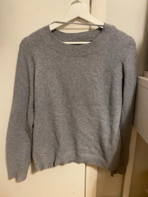 Grå stickad tröja med rund hals - Mysig grå stickad tröja med rund halsringning och ribbade muddar. Perfekt för lager på lager under kyliga dagar. Enkel och stilren design som passar till det mesta.