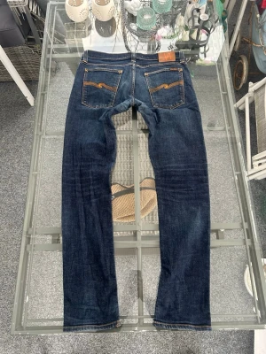 Nudie Jeans – Tight Long John (W34 L34) - Snygga Nudie Jeans Tight Long John i mörkblå indigo. Tight/slim passform med stretch som sitter clean på benen. Klassisk Nudie-design med signatur­sömmar på bakfickorna. Tillverkade i ekologisk bomull, perfekt för en minimalistisk och modern stil.  🧵 98% ekologisk bomull, 2% elastan 📏 W34 L34 👖 Tight / slim fit 🎨 Mörkblå denim 📌 Mycket bra skick  Passar perfekt till sneakers, boots eller loafers – easy everyday jeans. 