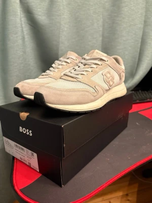 Beige sneakers från BOSS - Snygga sneakers från BOSS i beige och vitt med mesh och mockadetaljer. Skorna har rund tå, platt sula och snörning. BOSS-logga på sidan och plösen. Perfekta för dig som gillar stilrena och neutrala färger.