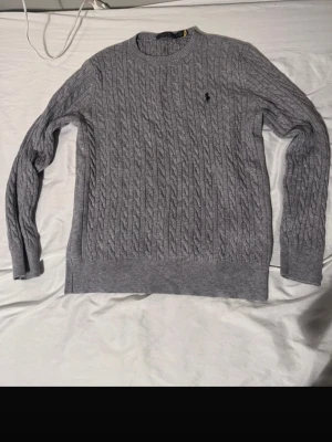 Grå kabelstickad tröja Polo Ralph Lauren - Säljer en grå kabelstickad tröja från Polo Ralph Lauren i storlek M. Tröjan har rund halsringning, långa ärmar och den klassiska broderade loggan på bröstet. Perfekt för dig som gillar stilrena och tidlösa plagg.