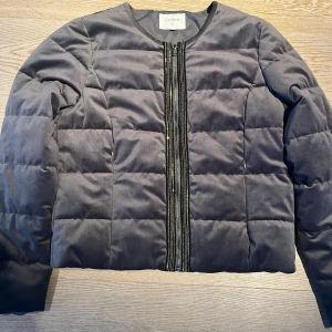 Grå quiltad pufferjacka från Cream - Snygg grå pufferjacka från Cream med quiltad design och mocka liknande finish. Jackan har rund halsringning och dragkedja framtill med dekorativa ränder i svart och guld. Perfekt för kyliga dagar och enkel att matcha med olika outfits. 