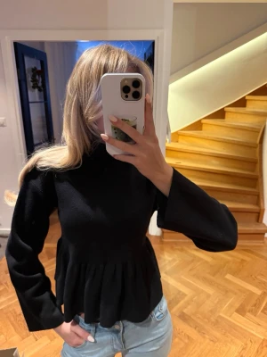 Svart blus med volang och polokrage - Säljer en svart blus med hög krage och långa ärmar. Toppen har en markerad midja och volang nedtill som ger en snygg siluett. Materialet känns mjukt och lite tjockare, perfekt för kyligare dagar. Enkel men ändå chic design som passar till jeans eller kjol.