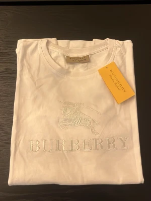 Vit Burberry t-shirt med broderi - Snygg vit t-shirt från Burberry i bomull med broderad logga och riddare framtill. Klassisk rund hals och korta ärmar. Perfekt för dig som gillar stilrena och exklusiva plagg med diskreta detaljer. Lite rosa färg som försvinner efter en tvätt vid storlekalappen 